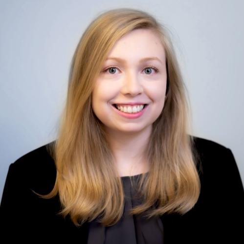 Rebecca has long blond hair. She is smiling and wearing a black jacket and purple blouse