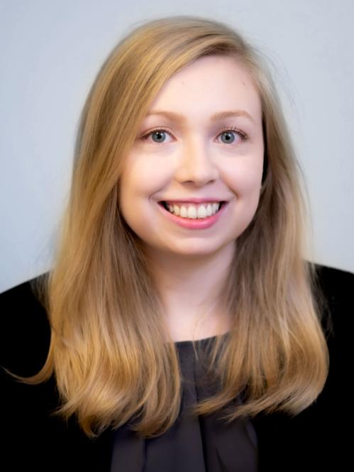 Rebecca has long blond hair. She is smiling and wearing a black jacket and purple blouse