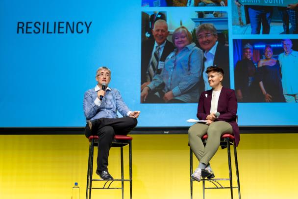 Two panelists sitting in chairs in front of a screen with images and the word resiliency