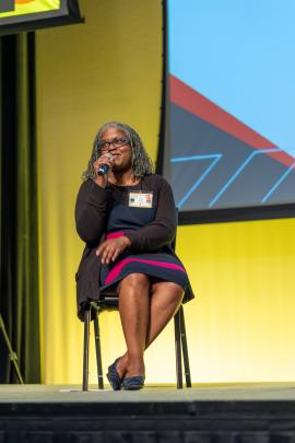A speaker seated on the stage speaking into a microphone