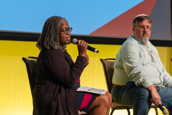 Two speakers seated on a stage, one on the left holding a microphone to speak